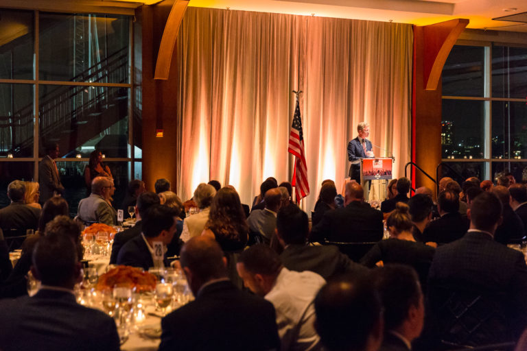 Speaker is on stage addressing the audience who is seating on round tables while dining