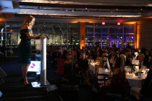 speaker on stage addressing the audience who is seating on dining tables.