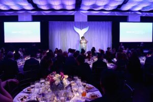guests eating dinner at round tables while speaker is on stage and she is presenting.