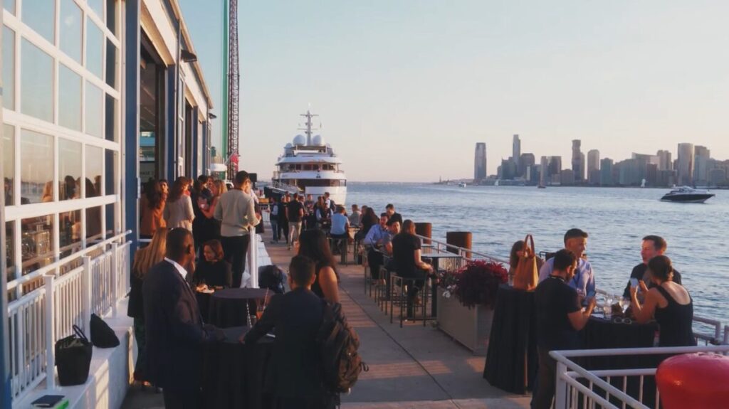 Guests at cocktail tables on the terrace beside a docked yacht at the Primary Summit at The Pier Sixty Collection