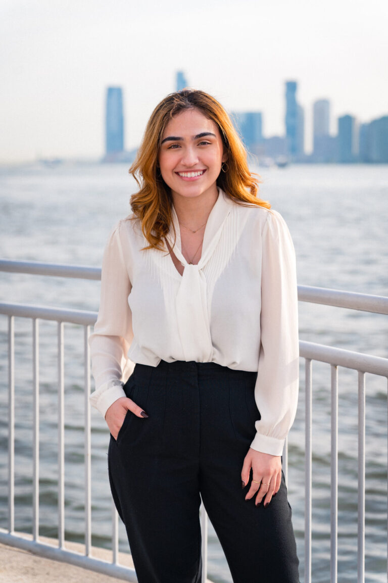 Jazmin Ayala smiling by the waterfront with the Manhattan skyline in the background at The Pier Sixty Collection