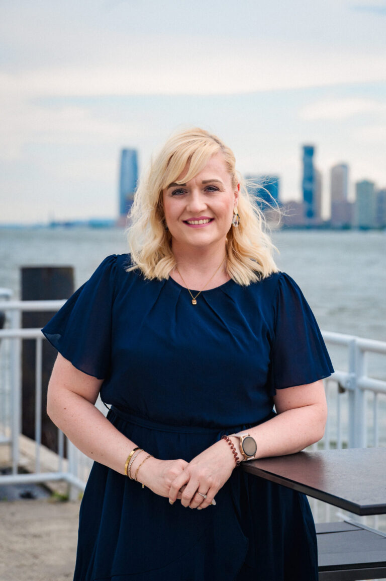 Stephanie Presume smiling by the waterfront with the Manhattan skyline in the background at The Pier Sixty Collection