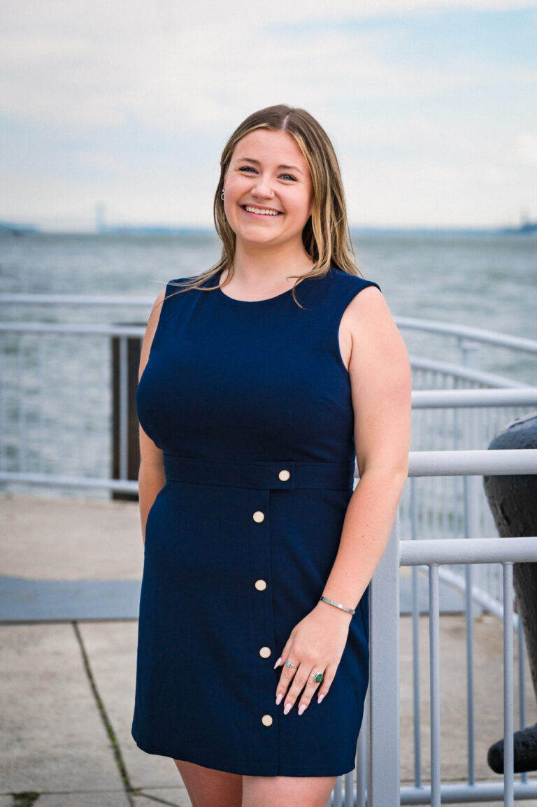 Annie Foley smiling by the waterfront with the Manhattan skyline in the background at The Pier Sixty Collection
