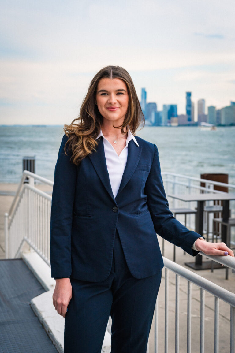 Faye Hogan smiling by the waterfront with the Manhattan skyline in the background at The Pier Sixty Collection